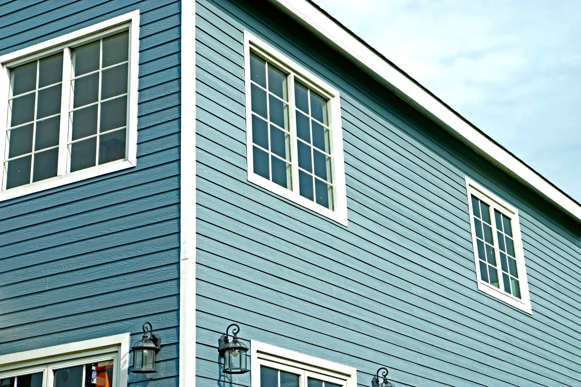 A close-up view of a blue house exterior with white trim in NJ, featuring three windows and two black metal lantern-style lights near the lower window. The sky is partly visible—an example of quality from a trusted Roofing Contractor Monmouth County.