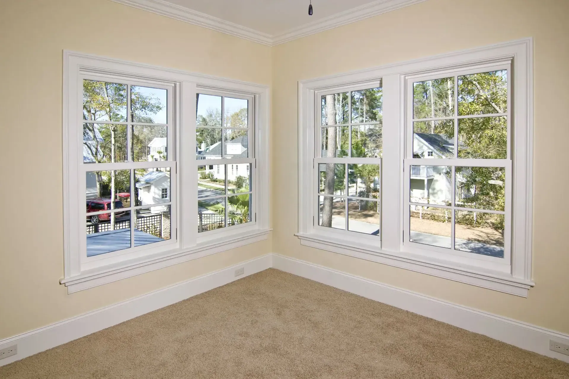 A bright, empty room with beige carpet, cream-colored walls, and large white-trimmed windows overlooking a suburban neighborhood—ideal for relaxing after a visit from your trusted Roofing Contractor Monmouth County, NJ.