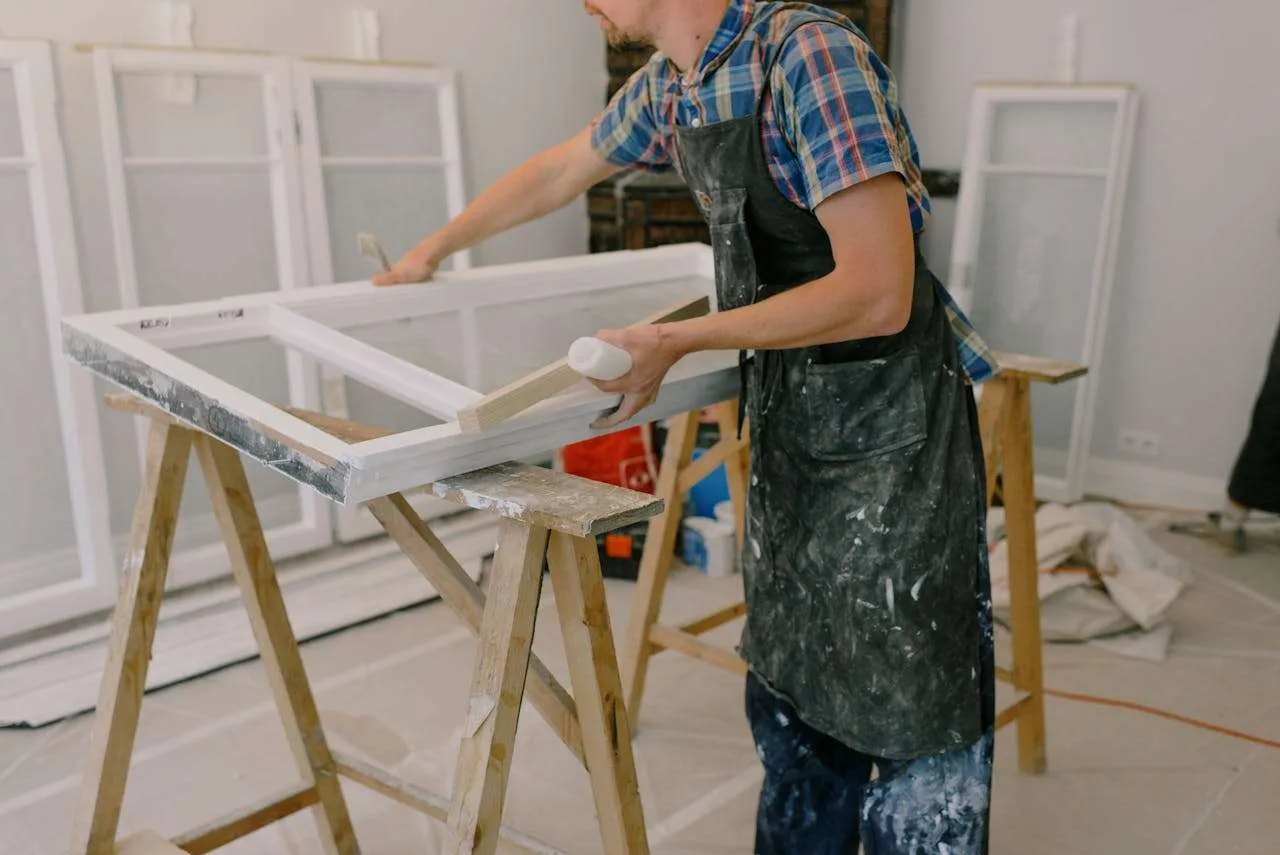 A person in a plaid shirt and paint-splattered apron is painting a window frame white, set on sawhorses in a workshop. Other frames suggest it's part of a NJ Roofing Contractor Monmouth County renovation project.