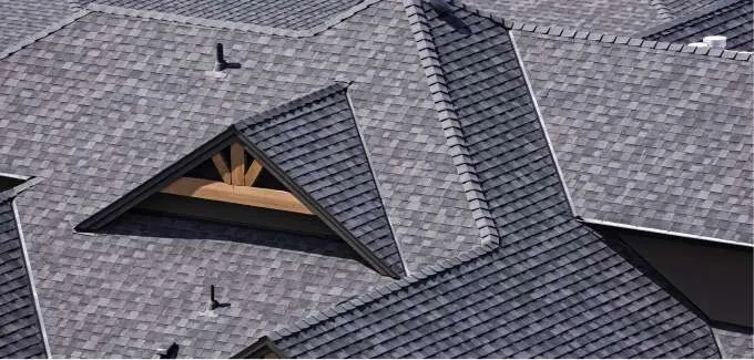 A close-up view of multiple intersecting gray shingled rooftops with various angles, ridges, and a triangular window showing wooden beams underneath—expertly crafted by a trusted roofing contractor in Monmouth County, NJ.