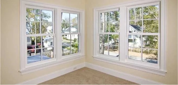 A bright corner of a room with beige walls and carpet, featuring two sets of large, white-trimmed windows that overlook a suburban NJ neighborhood with trees and houses outside. Ideal inspiration for any Roofing Contractor Monmouth County project.