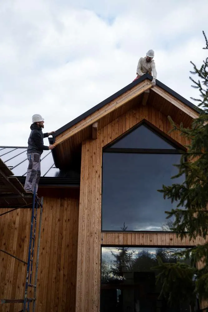 Two construction workers wearing safety helmets are installing or inspecting the roof of a wooden house—one on a ladder, the other atop the roof. A large window and tree complete this NJ scene for a trusted Roofing Contractor Monmouth County project.