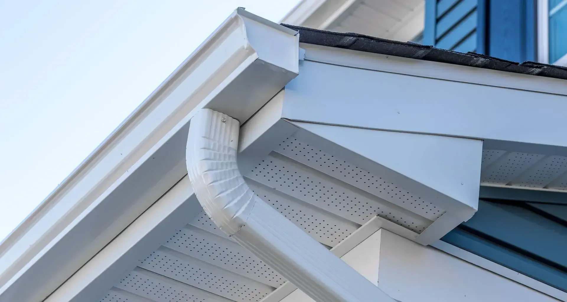 Close-up view of a white house roof corner, expertly maintained by a Roofing Contractor Monmouth County, NJ, showing a clean gutter system, downspout, soffit, and fascia against a clear sky with blue siding visible.