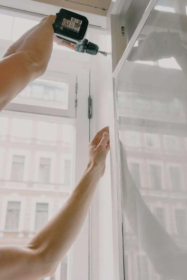 A person uses a power drill to install or fix a hinge on a white door or cabinet near a window with a city building visible outside, showcasing skills valued by a Roofing Contractor Monmouth County, NJ.