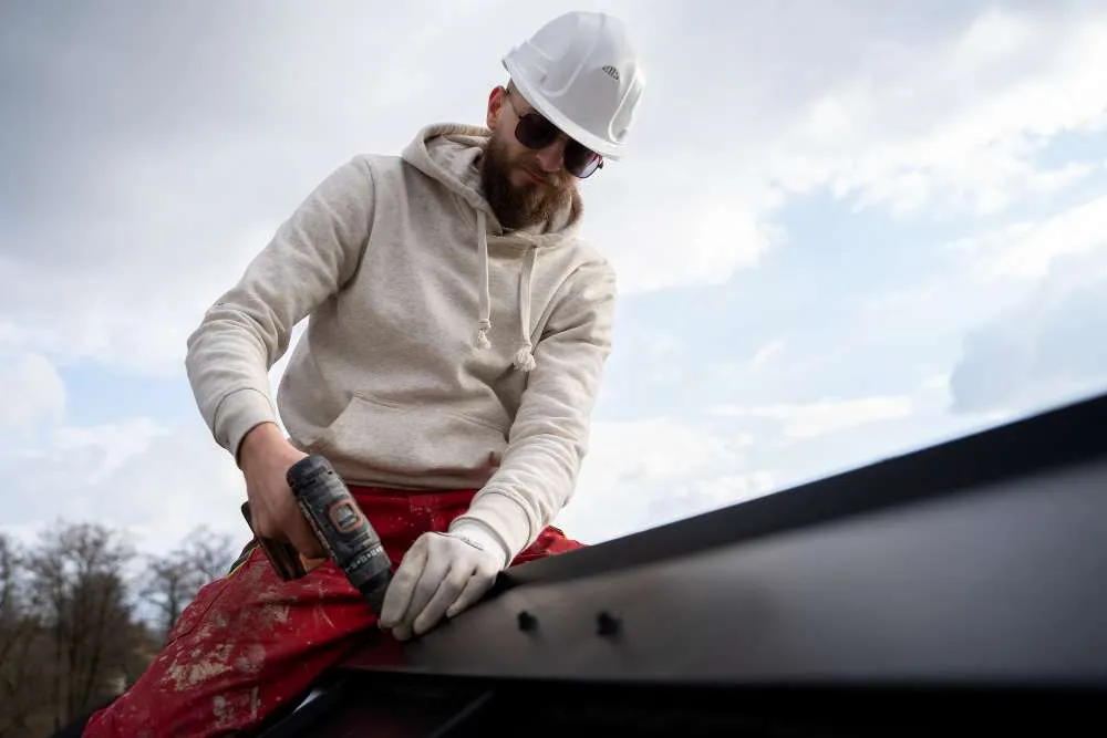 A construction worker in a white hard hat and safety glasses, working for an NJ roofing contractor in Monmouth County, uses a power drill to fasten screws into a metal beam outdoors under a partly cloudy sky.