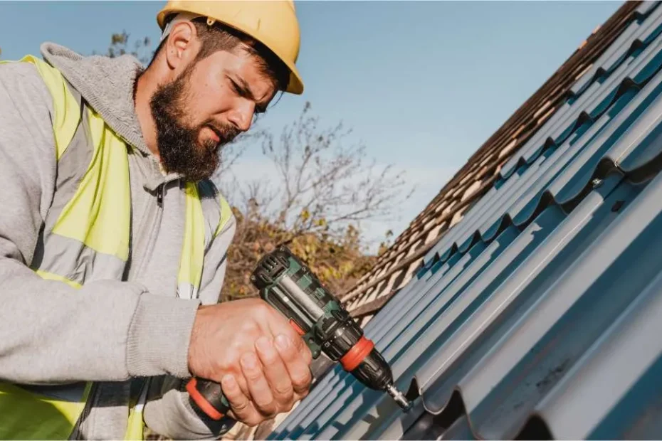 A construction worker in a yellow hard hat and reflective vest, employed by a Roofing Contractor Monmouth County NJ, uses a power drill to install metal roofing panels on a sloped roof outdoors under a clear sky.