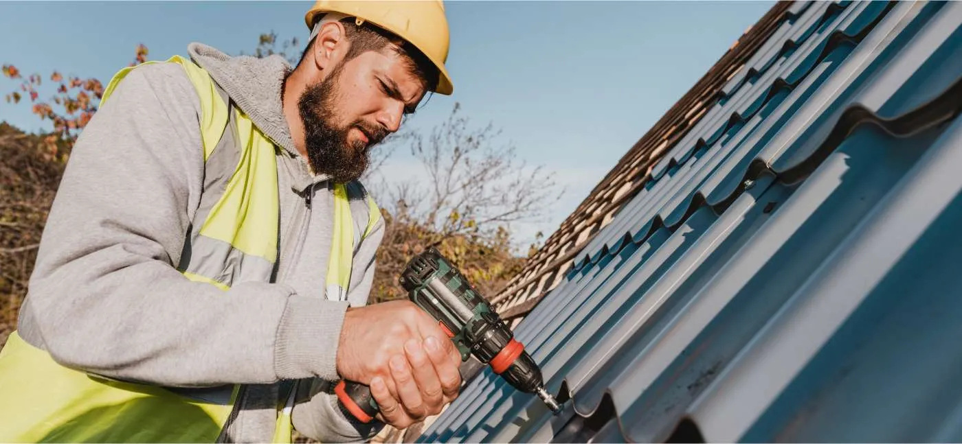 A construction worker in a yellow hard hat and reflective vest, employed by a Roofing Contractor Monmouth County NJ, uses a power drill to install metal roofing panels on a sloped roof outdoors under a clear sky.