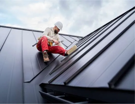 A construction worker wearing a white hard hat and red pants uses tools while sitting on a sloped metal roof under a cloudy sky, representing a dedicated Roofing Contractor Monmouth County, NJ.