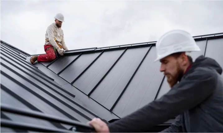 Two construction workers in hard hats are installing or repairing a metal roof on a building. A Roofing Contractor Monmouth County team member is close to the camera, slightly out of focus, while another works further away on the rooftop in NJ.