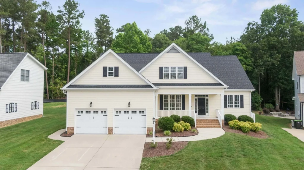 A modern two-story white house with a dark roof by a trusted Roofing Contractor Monmouth County, featuring a two-car garage, front porch, manicured lawn, and shrubs, set against tall trees and neighboring homes in NJ.
