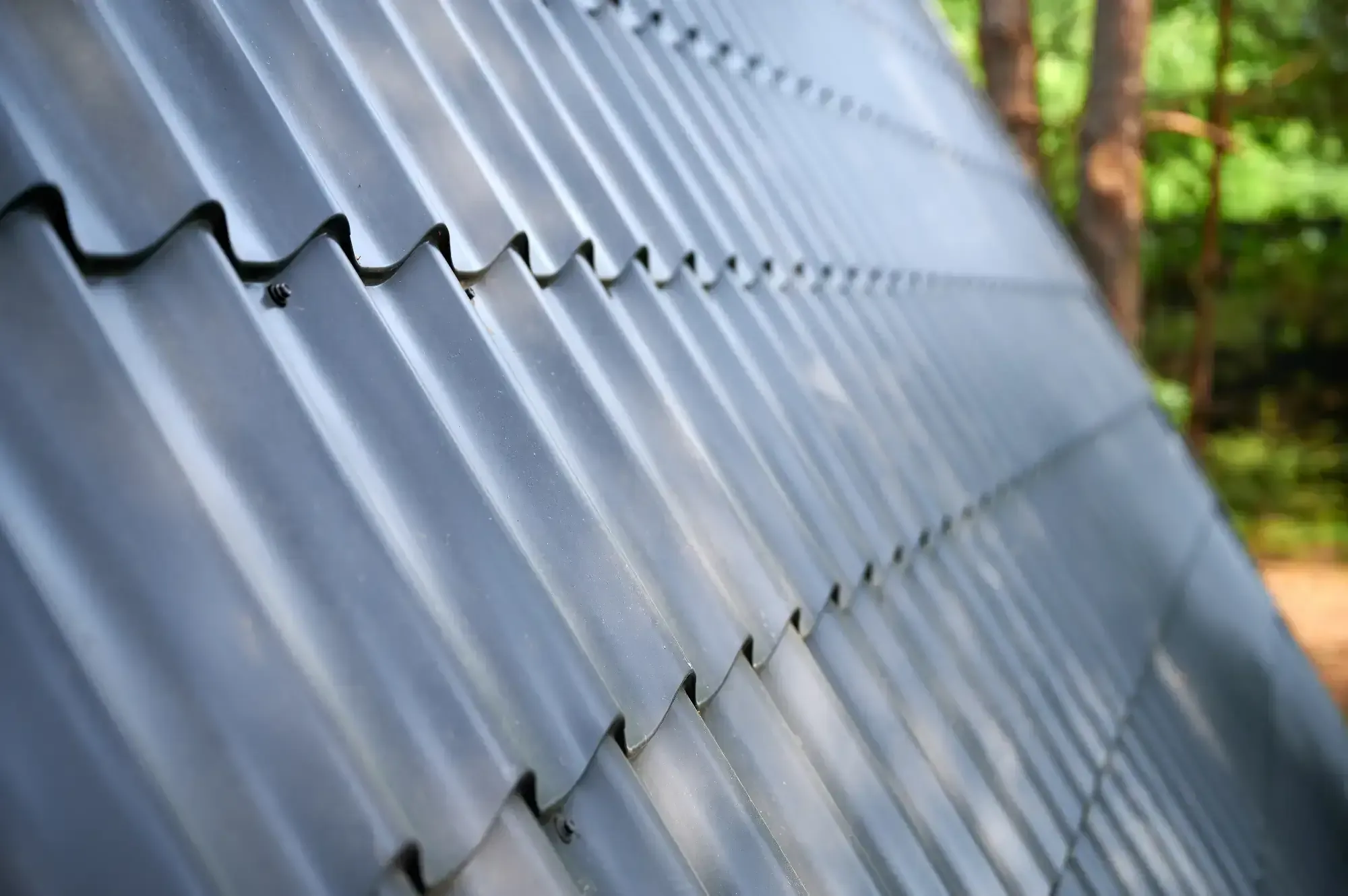 Close-up view of a metal sheet roof installed by a Roofing Contractor Monmouth County, featuring overlapping panels and wavy ridges. Trees and greenery are visible out of focus in the NJ background.