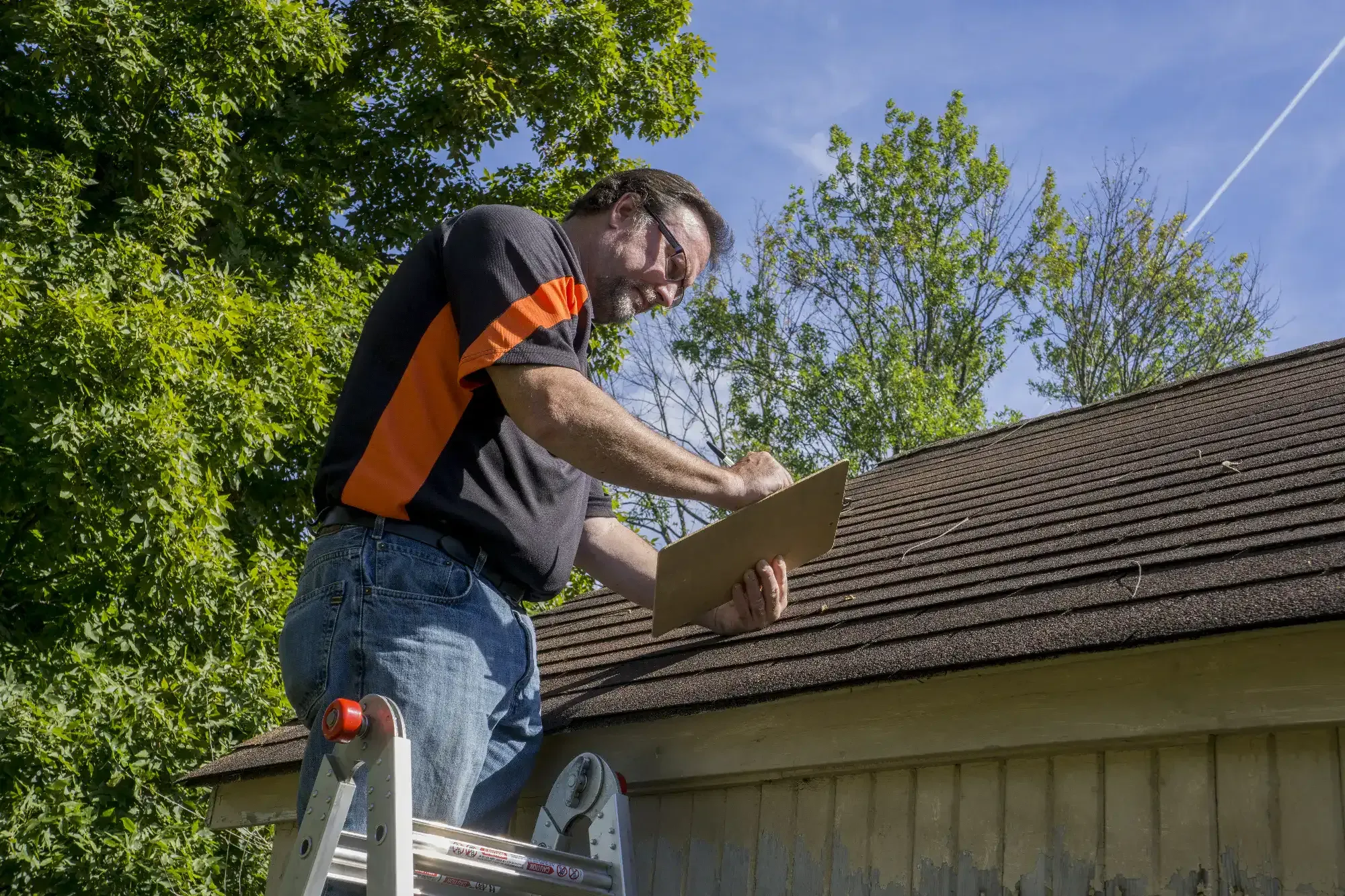 A Roofing Contractor in Monmouth County, NJ stands on a ladder inspecting a roof, writing notes on a clipboard. He wears safety glasses, a black and orange shirt, and jeans, with trees and blue sky in the background.