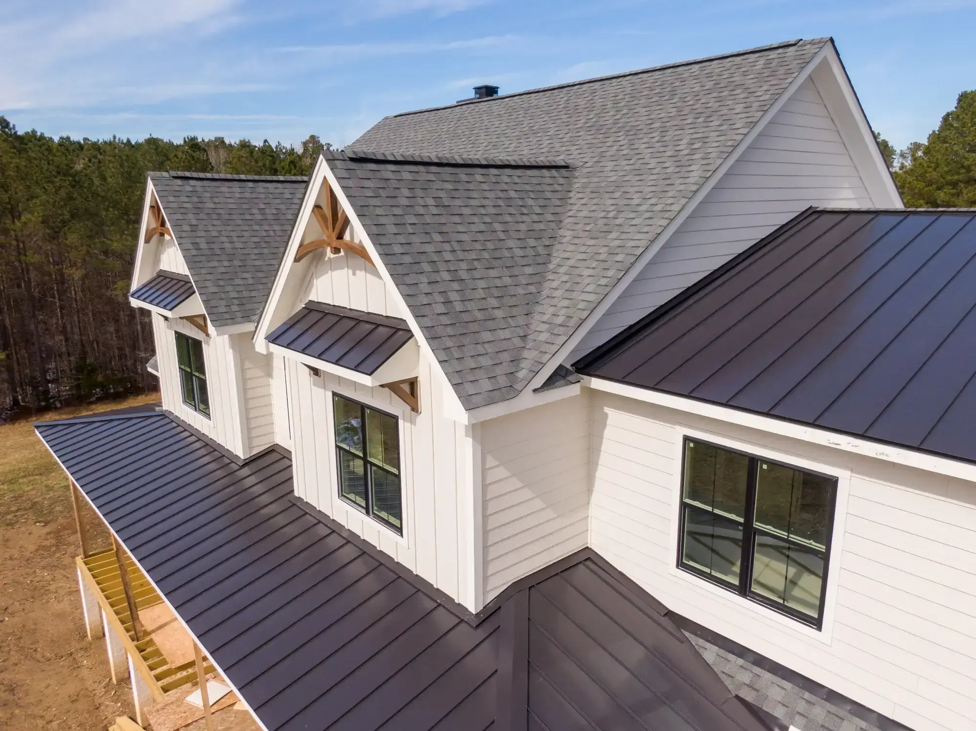 A modern farmhouse in NJ with white siding, black framed windows, and a mix of gray asphalt shingles and black metal roofing by a trusted Roofing Contractor Monmouth County, surrounded by trees under a blue sky.