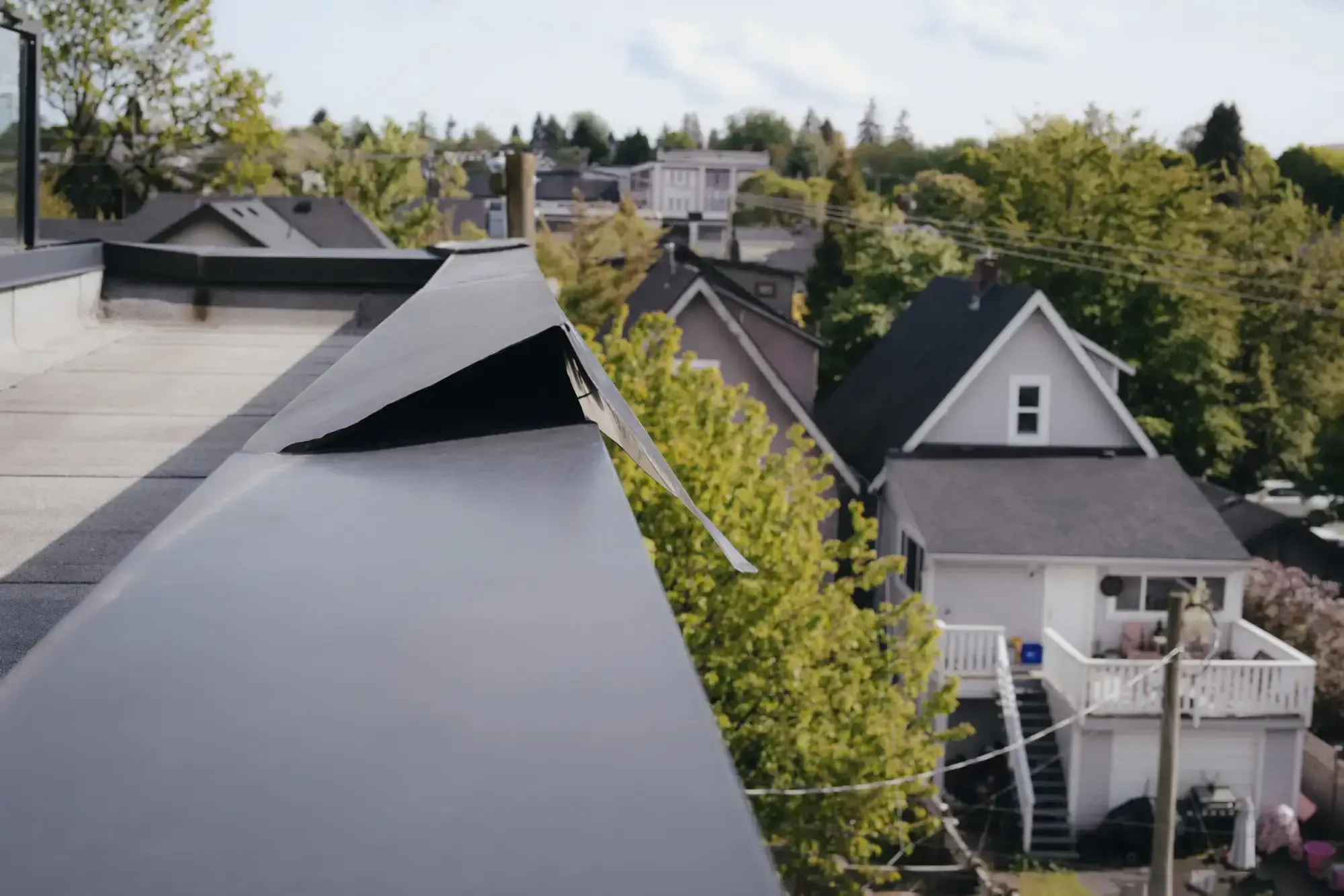 A close-up of a metal roof edge with a piece of flashing bent upwards, overlooking residential houses and trees in a suburban neighborhood&mdash;expertly detailed by a trusted Roofing Contractor in Monmouth County, NJ.