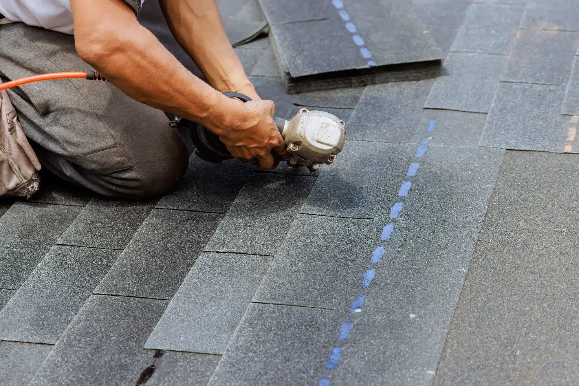 A Roofing Contractor Monmouth County kneels on a roof in NJ, installing asphalt shingles with a nail gun. Several shingle pieces and a dotted blue guideline are visible on the roof surface.
