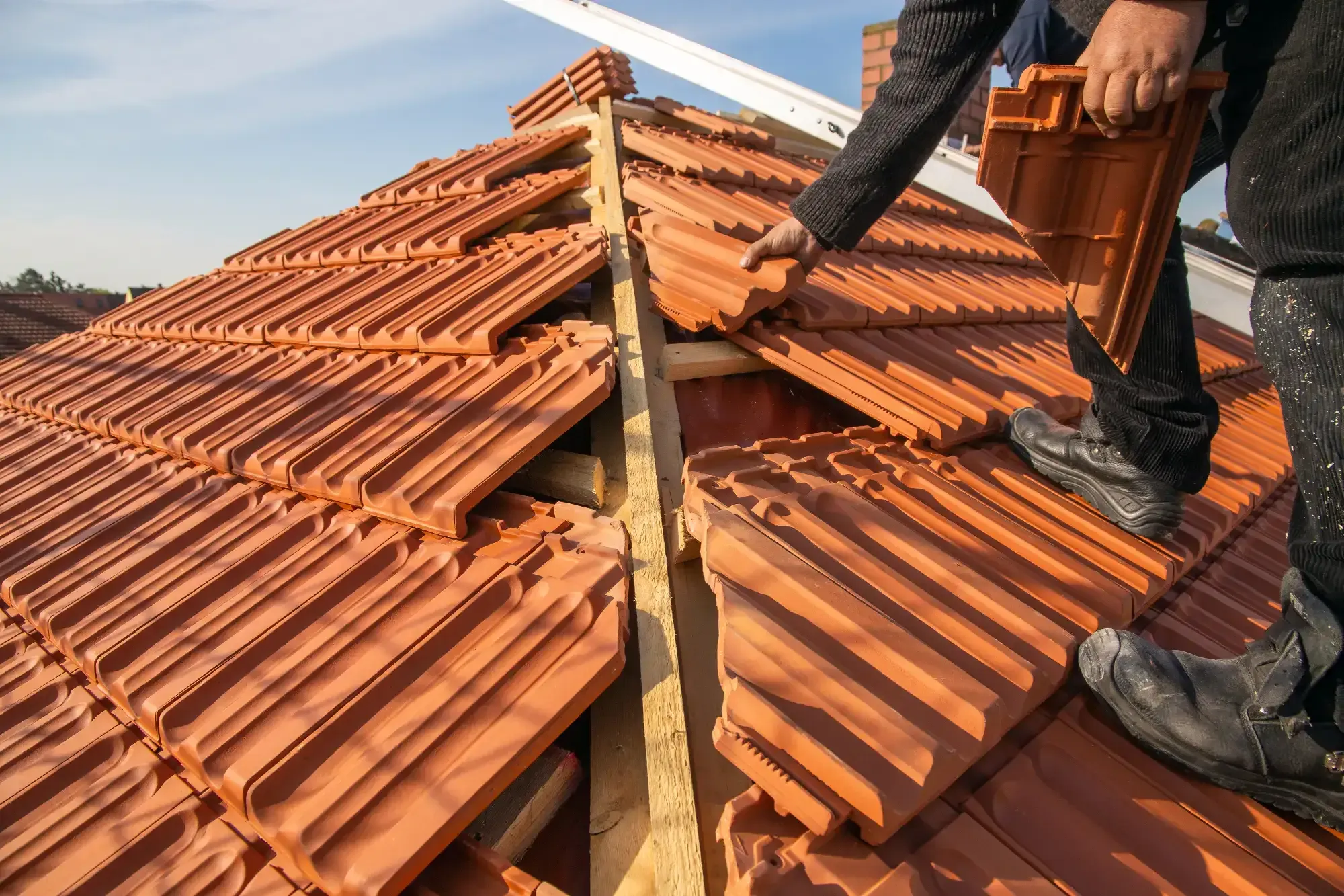 A roofing contractor in Monmouth County, NJ installs orange clay roof tiles on a wooden framework, carefully placing a tile near the roof&rsquo;s ridge under clear daylight.