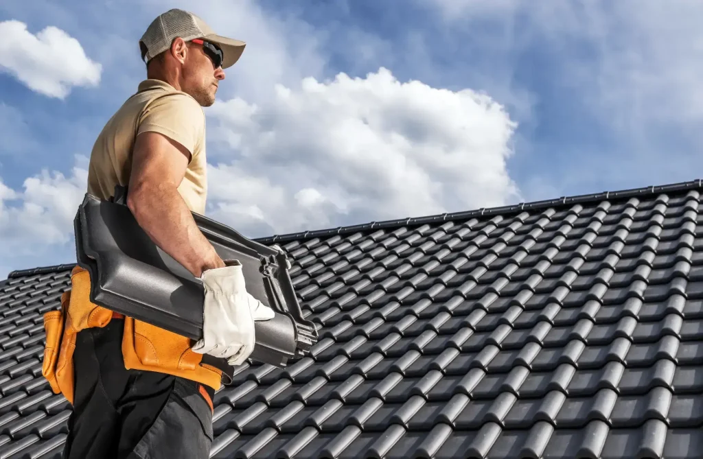 A worker wearing a cap, sunglasses, gloves, and a tool belt carries roof tiles beside a modern NJ roof with dark, shiny tiles under a partly cloudy sky—representing skilled Roofing Contractor Monmouth County professionals at work.