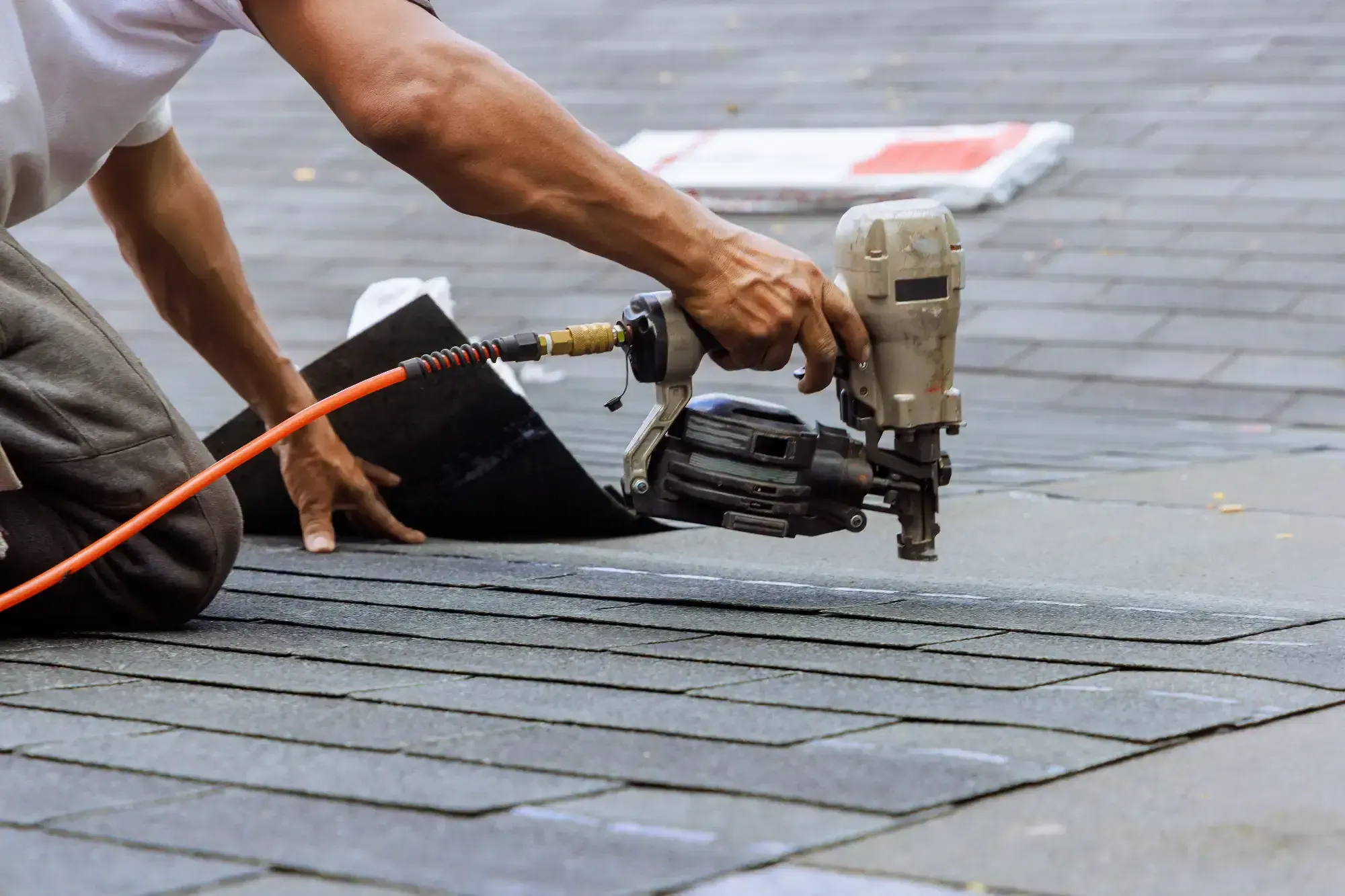 A Roofing Contractor Monmouth County kneels on a gray shingled roof in NJ, using a pneumatic nail gun with both hands to secure asphalt shingles during installation or repair.