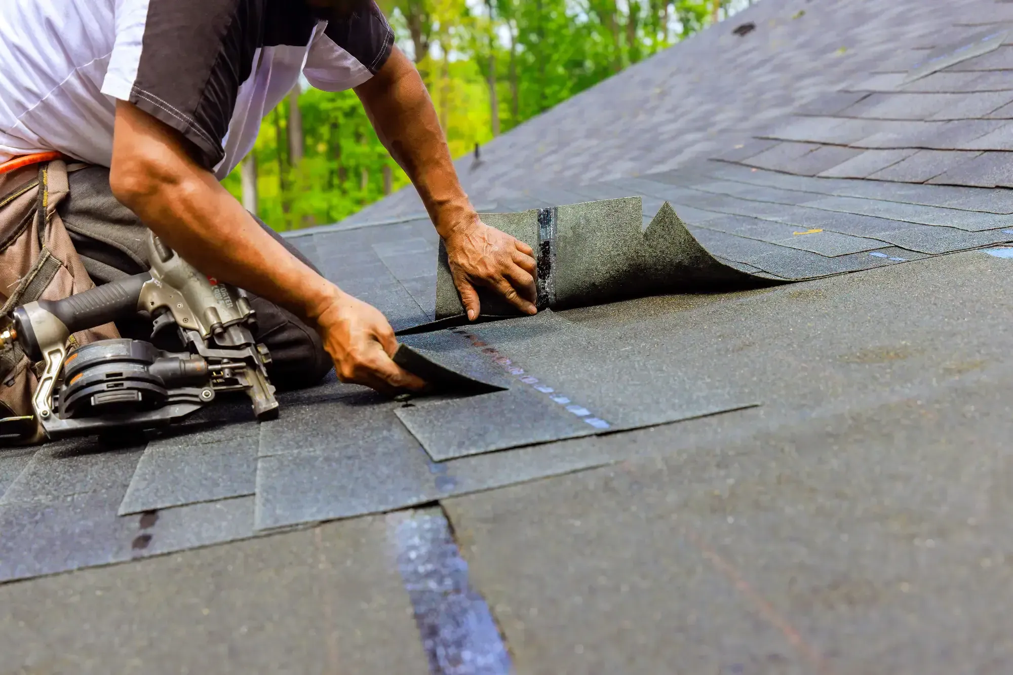 A Roofing Contractor in Monmouth County, NJ is installing asphalt shingles by hand, with a roofing tool placed nearby. Green trees are visible in the blurred background.