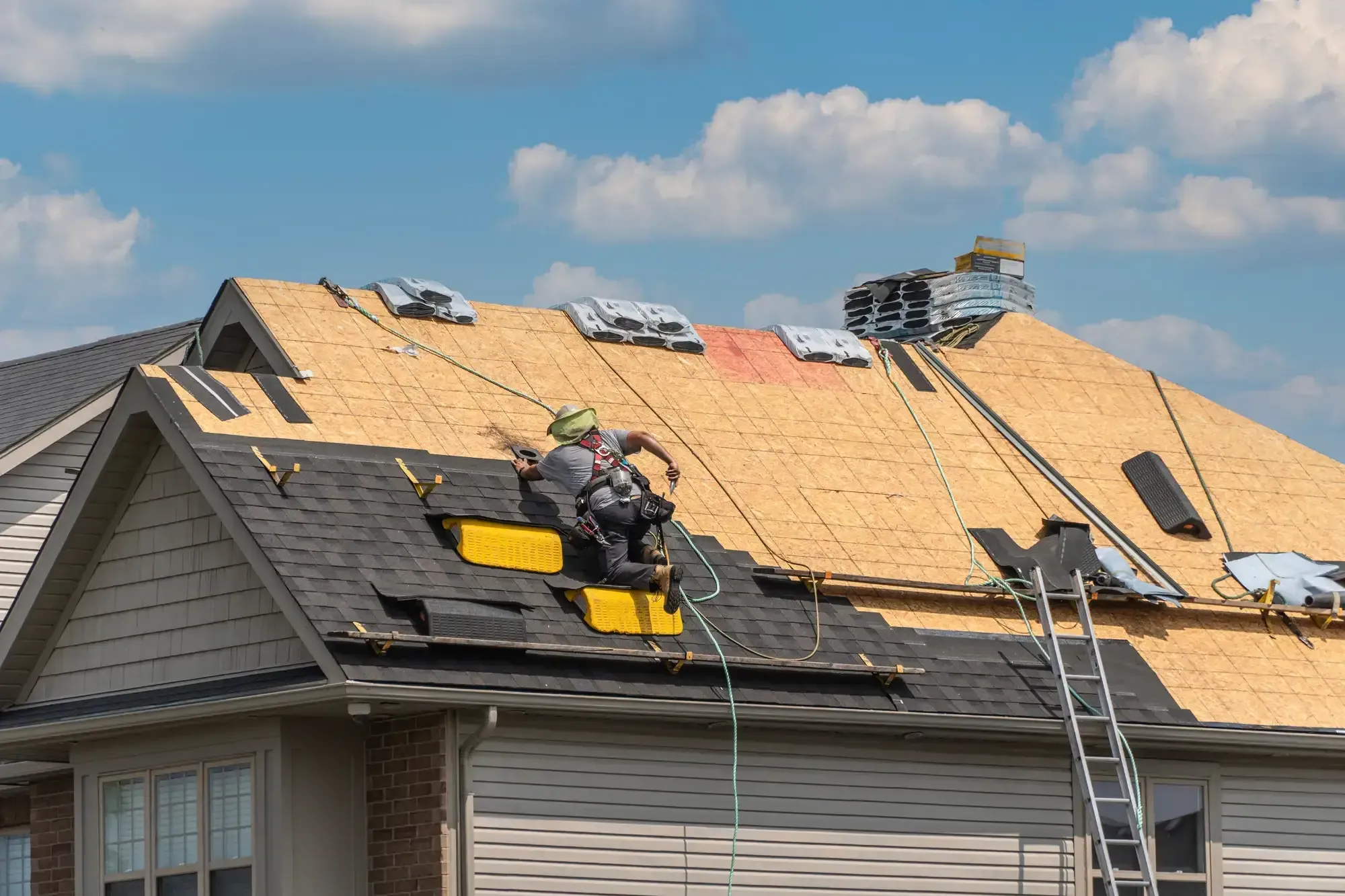 A Roofing Contractor in Monmouth County, NJ, wearing safety gear, repairs a house roof by installing new shingles on wooden sheathing. Construction materials and tools are spread out, with a ladder propped against the house under a blue sky.