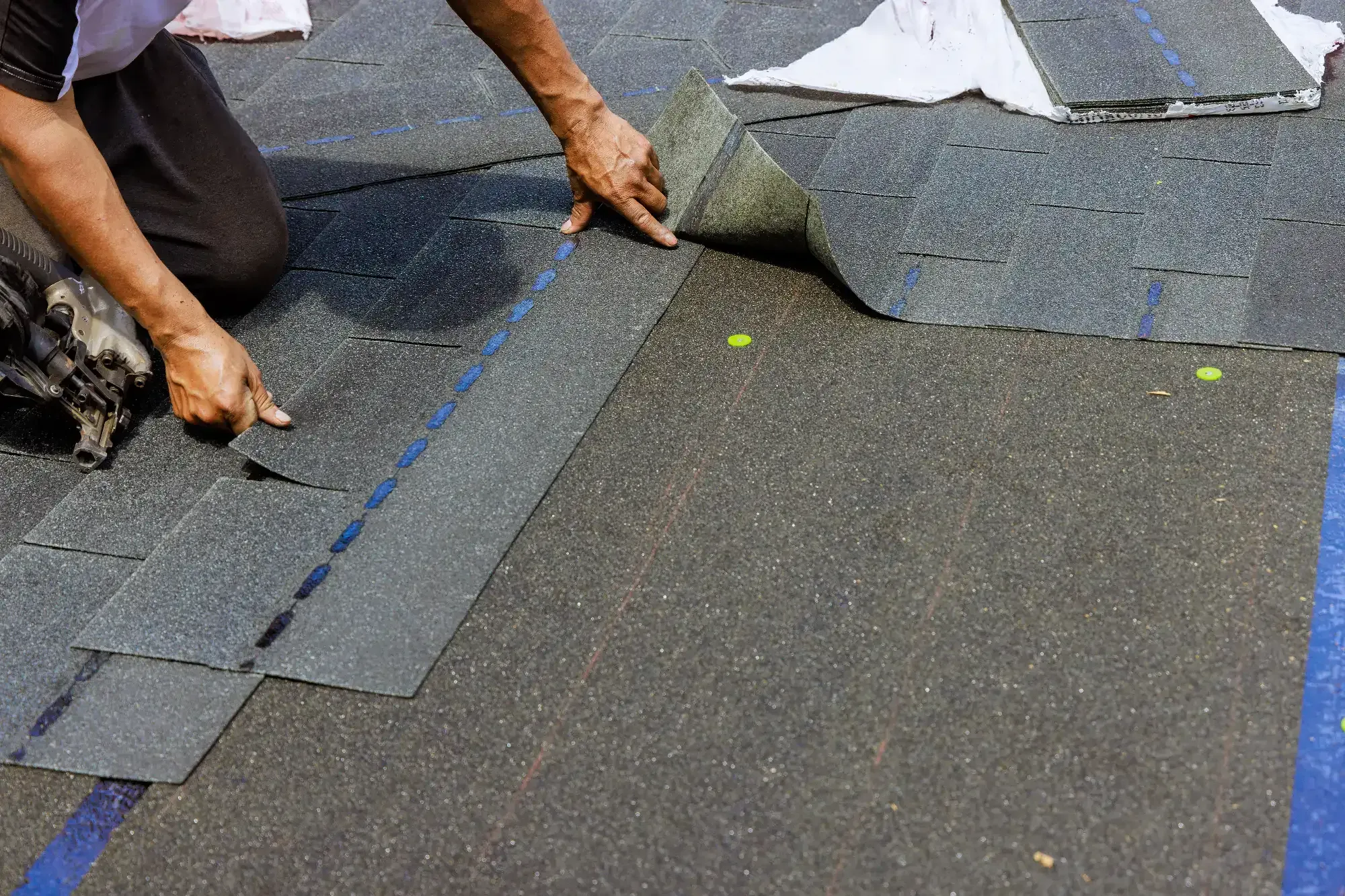 A Roofing Contractor Monmouth County installs asphalt roof shingles by hand, carefully positioning each shingle while other roofing materials and tools are visible on the NJ rooftop.
