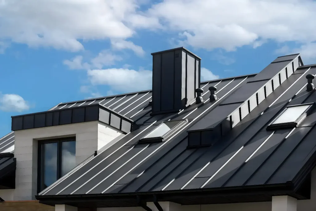 A modern house in NJ with a dark metal roof featuring angled skylights, a large vertical chimney, and several small roof vents—showcasing the work of a top Roofing Contractor Monmouth County—set against a blue sky with scattered clouds.