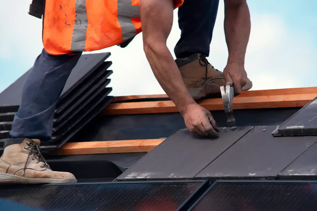 A Roofing Contractor in Monmouth County, NJ, wearing an orange safety vest, uses a hammer to install roof tiles. Several stacks of tiles and wooden beams are visible, along with part of the worker's boots.