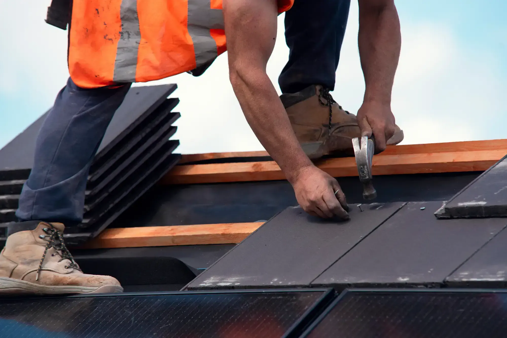 A Roofing Contractor in Monmouth County, NJ, wearing an orange safety vest, uses a hammer to install roof tiles. Several stacks of tiles and wooden beams are visible, along with part of the worker's boots.