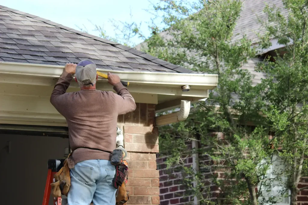 A Roofing Contractor in Monmouth County, NJ, in work clothes uses a level tool to check the alignment of a rain gutter on the roof edge of a brick house, standing on a ladder near some trees.