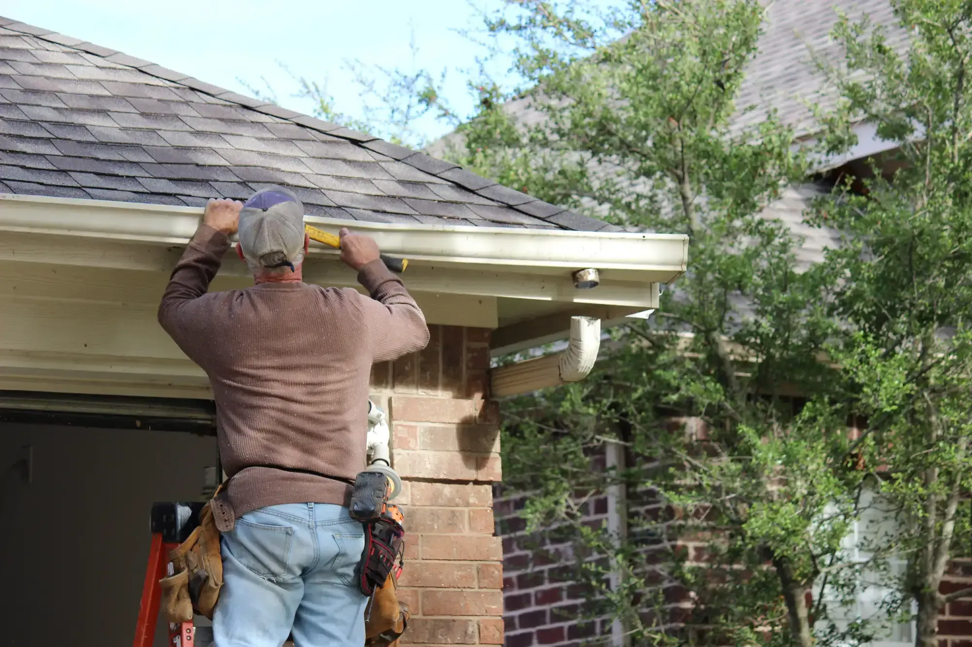 A Roofing Contractor in Monmouth County, NJ, in work clothes uses a level tool to check the alignment of a rain gutter on the roof edge of a brick house, standing on a ladder near some trees.