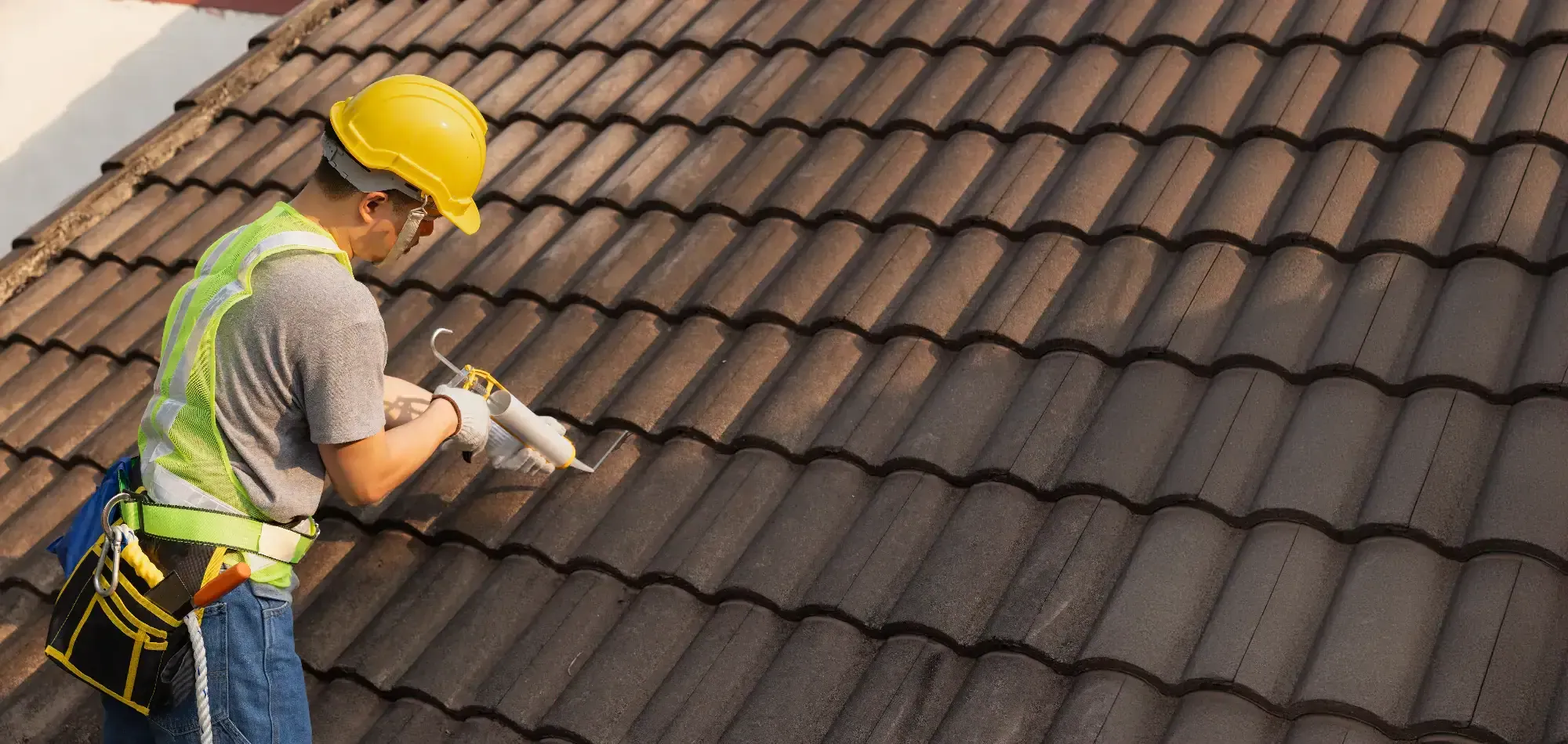 A Roofing Contractor in Monmouth County, NJ, wearing a yellow hard hat and safety gear, applies sealant to brown roof tiles while working on a sloped roof.