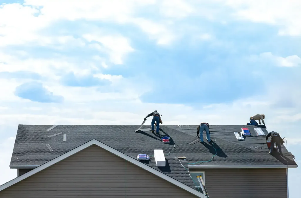 Four workers from a Roofing Contractor Monmouth County, NJ, are installing new shingles on the roof of a house under a partly cloudy sky, with roofing materials and tools scattered across the roof.
