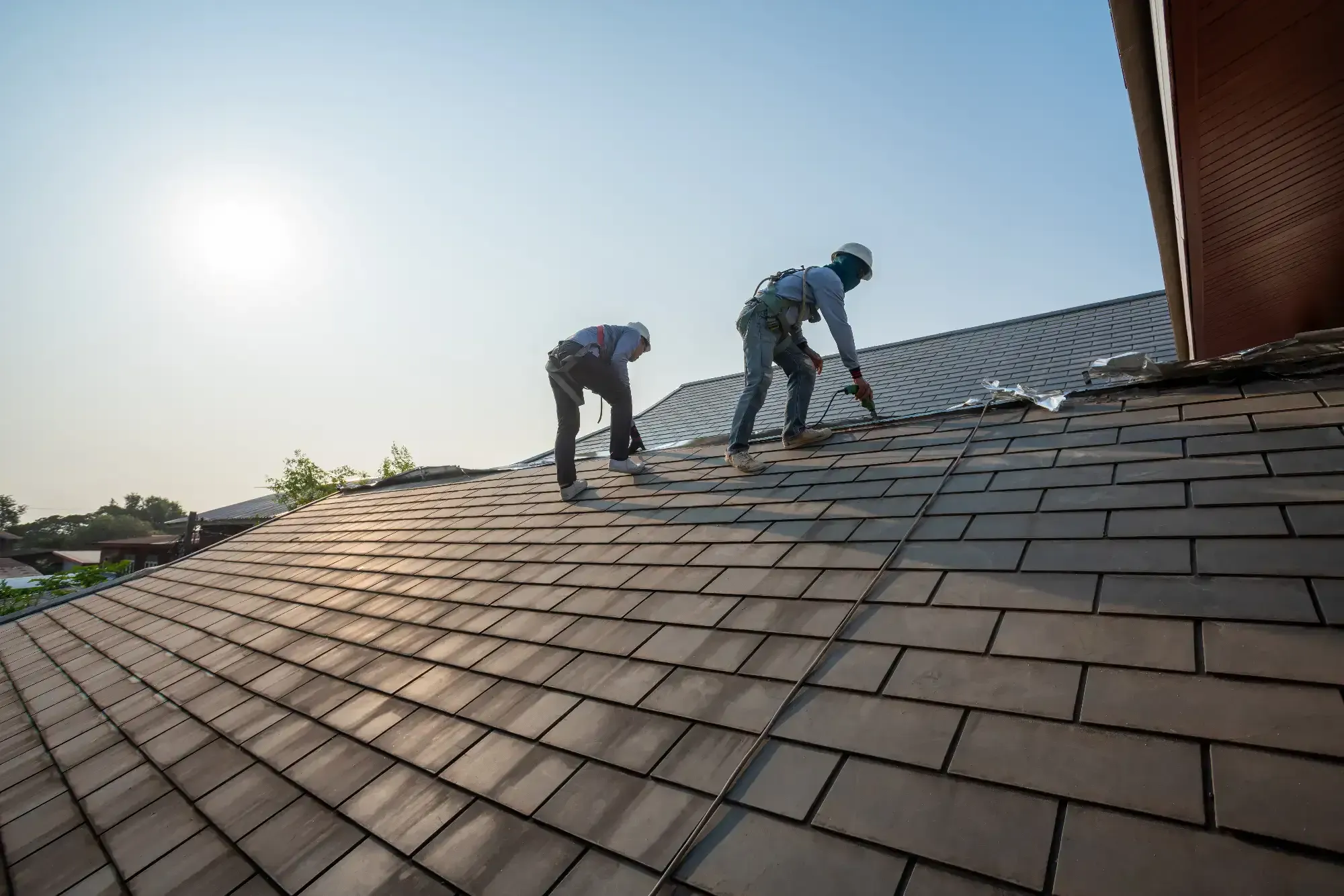 Two workers wearing safety gear are installing or repairing shingles on a sloped roof under bright sunlight, with houses and trees in the background&mdash;a typical scene for a Roofing Contractor Monmouth County, NJ.