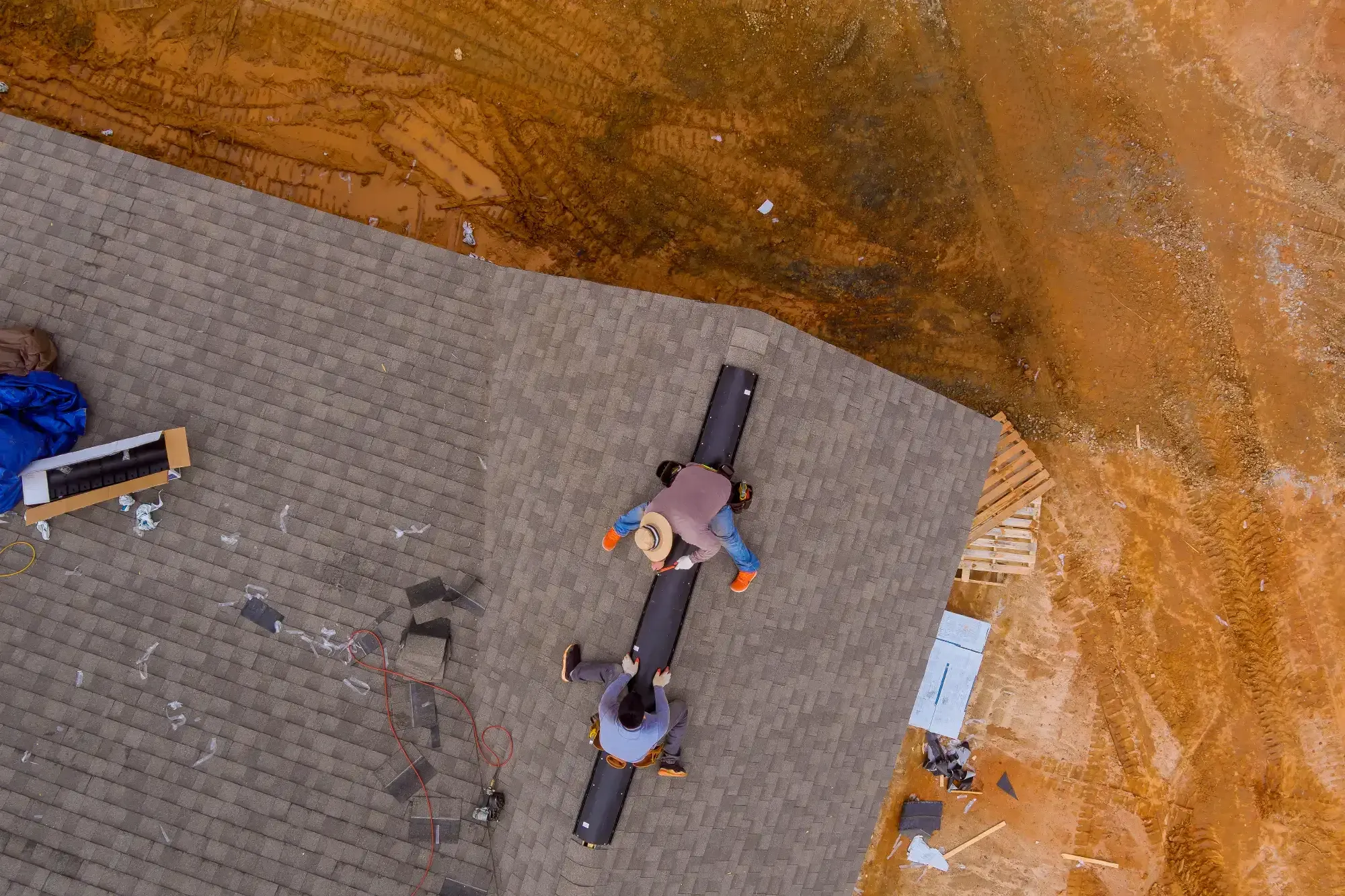Aerial view of Roofing Contractor Monmouth County, NJ workers installing shingles on a house roof at a construction site, with tools and equipment scattered nearby on the roof and ground.