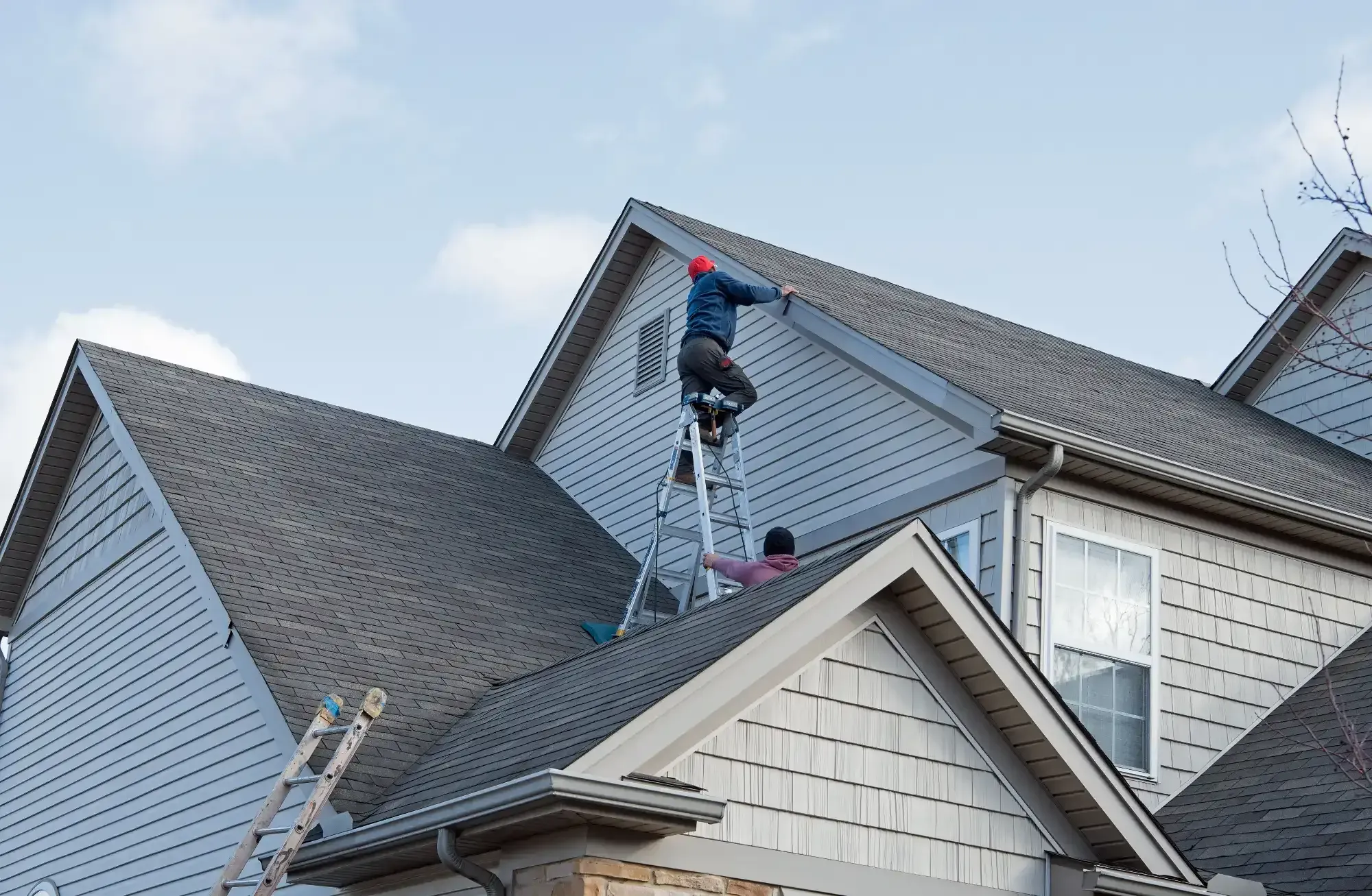 Two people work on the roof of a house; one stands on a ladder placed on the roof, while the other is below on a lower section. Another ladder leans against the house&mdash;typical scenes for an NJ Roofing Contractor Monmouth County trusts.