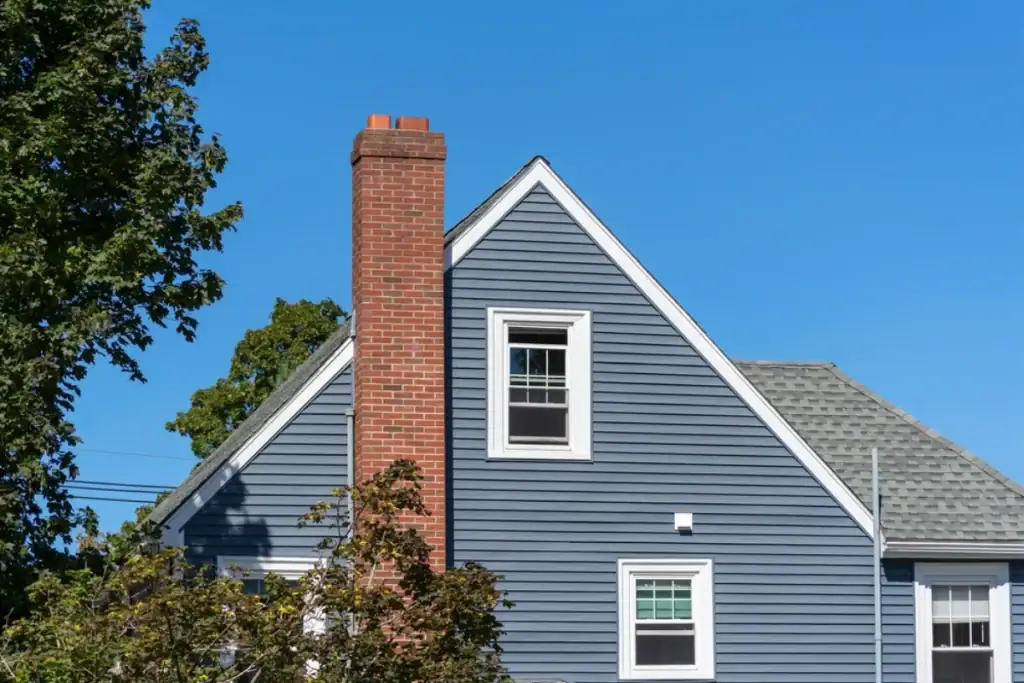 A blue house with white trim, a tall red brick chimney, and a steep roof—styled by a top Roofing Contractor Monmouth County—sits under a clear NJ sky, with green trees partially visible in the foreground.