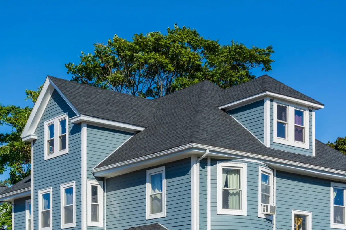 A close-up view of the upper part of a blue two-story house with white trim, several windows, and a dark shingle roof—expertly installed by a Roofing Contractor Monmouth County, NJ—framed by green trees and a clear blue sky.