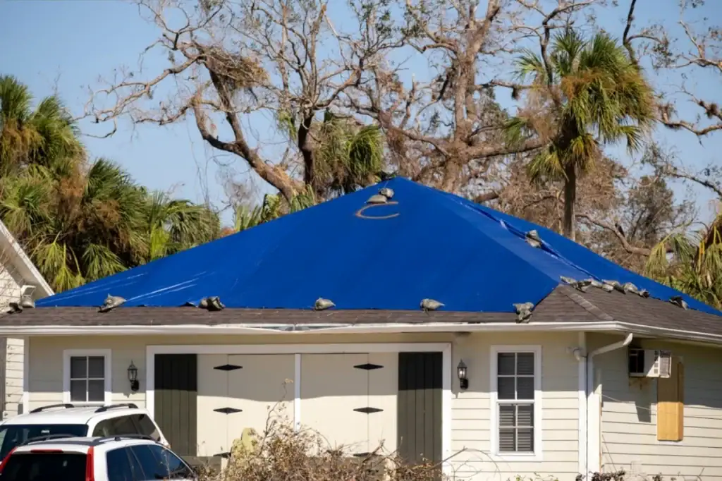A house with a blue tarp covering its roof, held down by sandbags, shows visible storm damage. In the background, leafless and broken trees reveal recent severe weather—an urgent situation for a roofing contractor in Monmouth County, NJ.