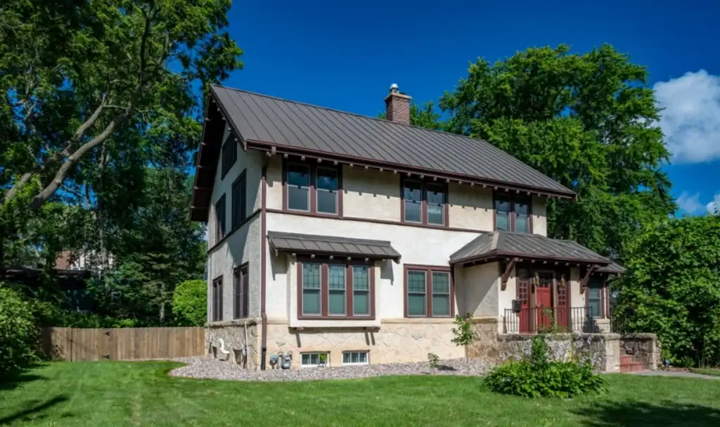 A two-story house with cream-colored stucco walls, dark brown trim, and a dark metal roof installed by a trusted roofing contractor in Monmouth County, NJ, surrounded by green grass, trees, and a wooden fence under a bright blue sky.