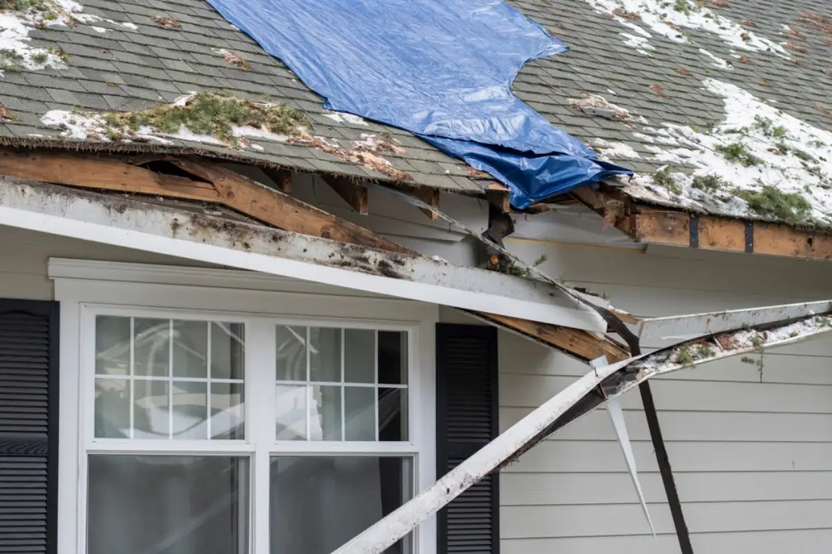 A house in NJ with visible roof damage, broken beams, and part of the roof covered by a blue tarp. Snow and debris rest above a white-framed window with black shutters—perfect for a Roofing Contractor Monmouth County to repair.