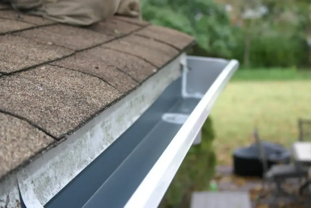 A close-up view of a house roof with clean, empty rain gutters along the edge; part of a person's legs are visible on the roof, suggesting someone—perhaps an NJ roofing contractor from Monmouth County—is cleaning or inspecting the gutters.