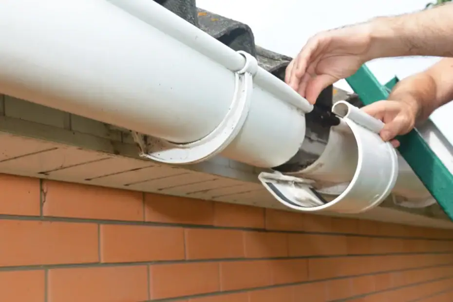 A Roofing Contractor Monmouth County professional repairs a white plastic rain gutter on a brick house in NJ, fixing a loose gutter bracket while standing on a green ladder.
