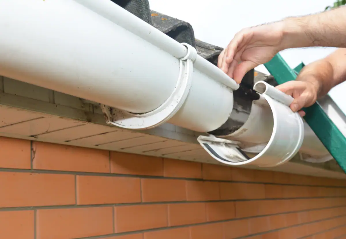 A Roofing Contractor Monmouth County professional repairs a white plastic rain gutter on a brick house in NJ, fixing a loose gutter bracket while standing on a green ladder.