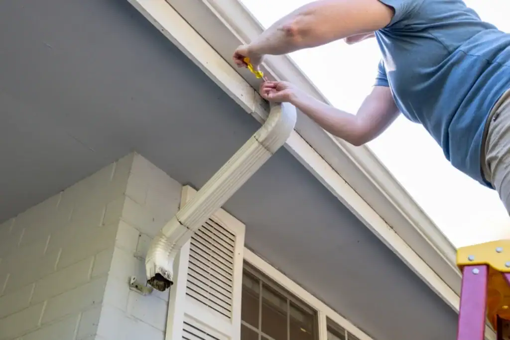 A person stands on a ladder using a tool to repair or install a white rain gutter downspout on the side of a house, assisted by an expert Roofing Contractor Monmouth County, NJ.