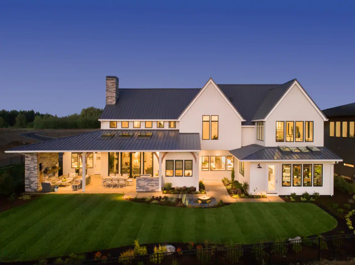 A large modern farmhouse with white siding, tall windows, and a gray metal roof—crafted by a top Roofing Contractor Monmouth County, NJ—is brightly lit at dusk. It features a spacious covered patio, stone chimney, and well-manicured landscaping.