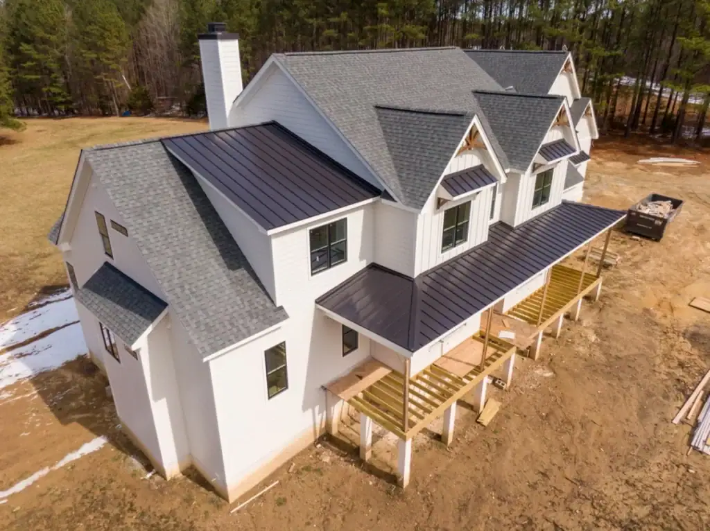 Aerial view of a large white house under construction, featuring gray shingle and black metal roofs by a top Roofing Contractor Monmouth County, NJ. The wooden porch frame is being built, with dirt and patches of grass and snow surrounding the site.