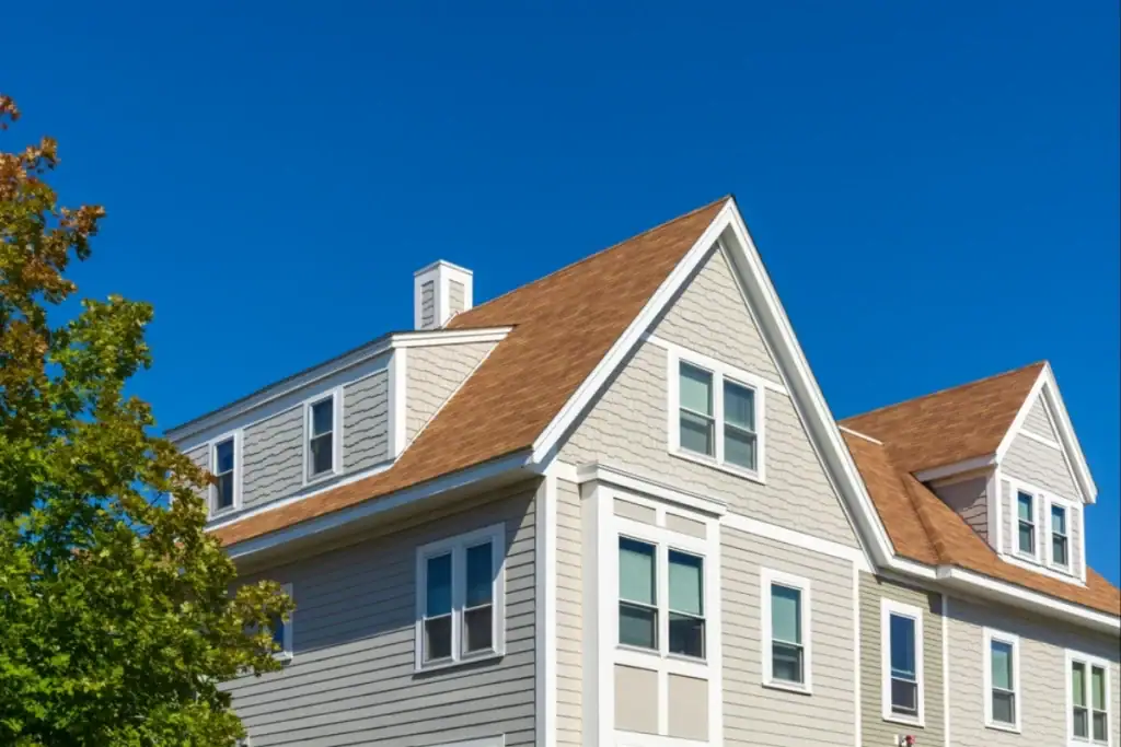A modern two-story house with beige siding, white trim, and a brown shingle roof is shown under a clear blue NJ sky, with part of a leafy green tree visible on the left side. Roofing Contractor Monmouth County services may have enhanced its curb appeal.