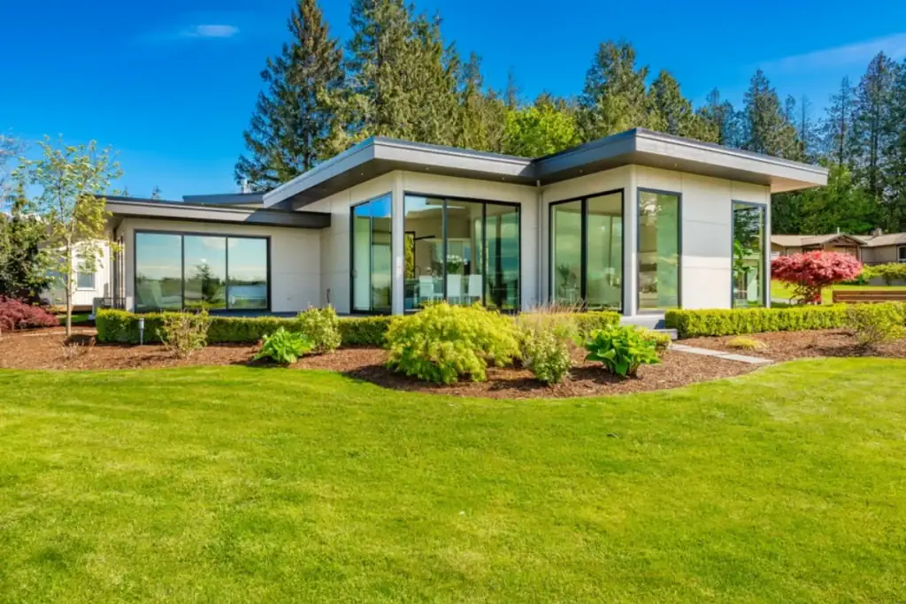A modern single-story house in NJ with large glass windows, flat roof, and white exterior, surrounded by a well-maintained green lawn and tall trees—a showcase for any top Roofing Contractor Monmouth County has to offer.