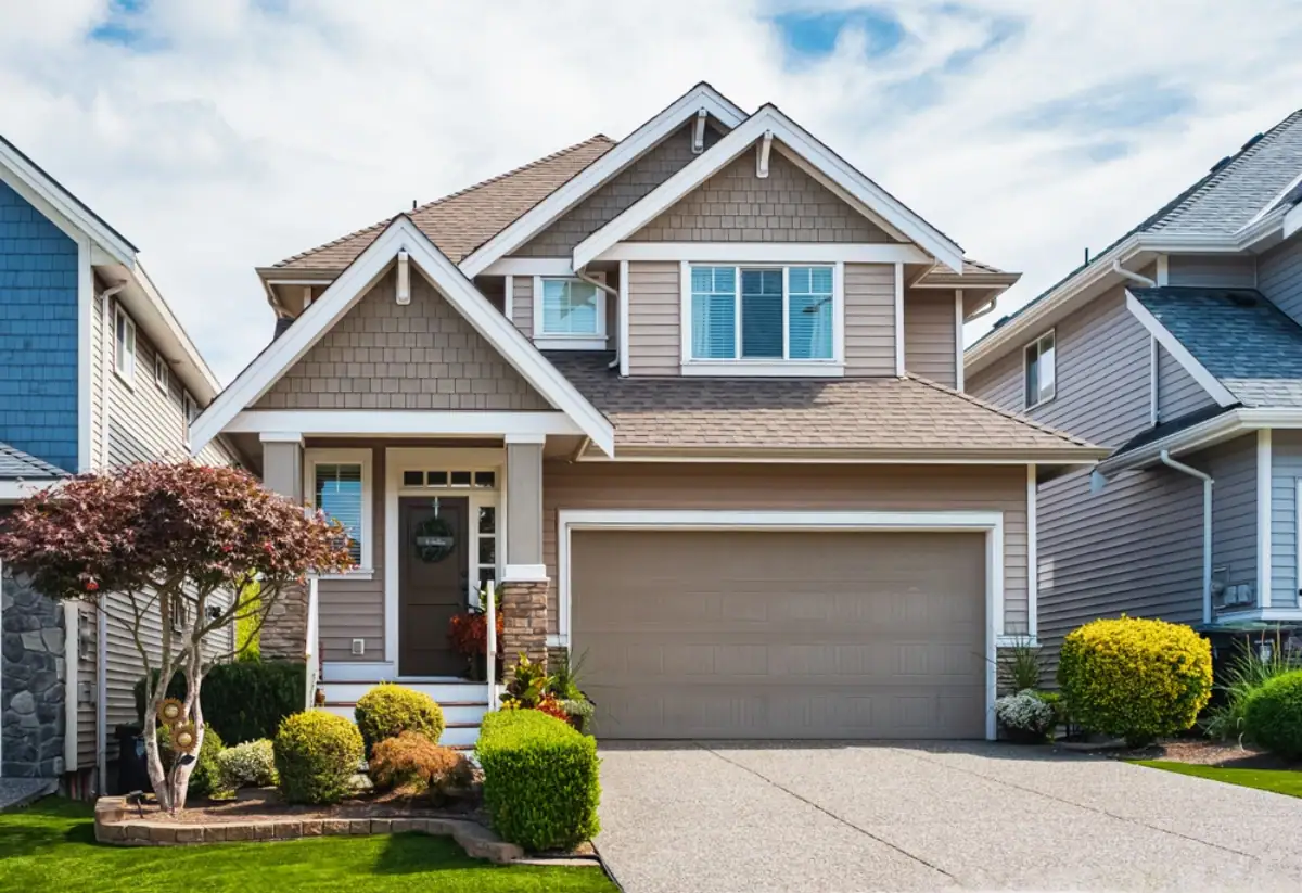Two-story suburban house in NJ with taupe siding, white trim, and a double garage. The front yard features green grass, neatly trimmed bushes, a small tree, and a concrete driveway. Trusted Roofing Contractor Monmouth County recommended.