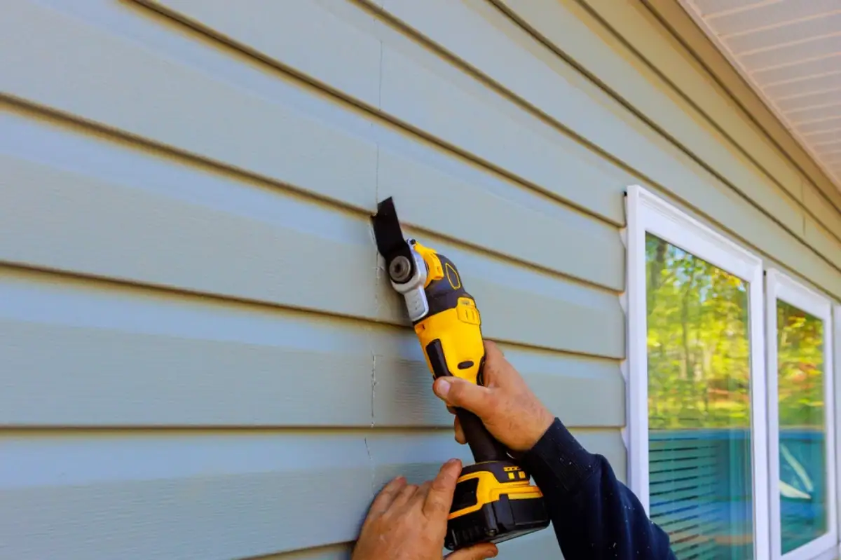 A Roofing Contractor in Monmouth County, NJ uses a yellow oscillating multi-tool to cut or trim blue vinyl siding on the exterior wall of a house, working precisely near a window.